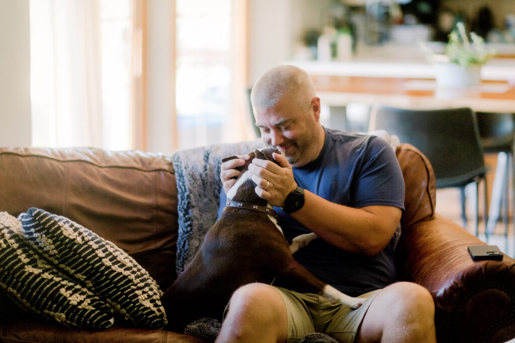 Man sitting on his couch at home while he pats and cuddles his brown and white dog