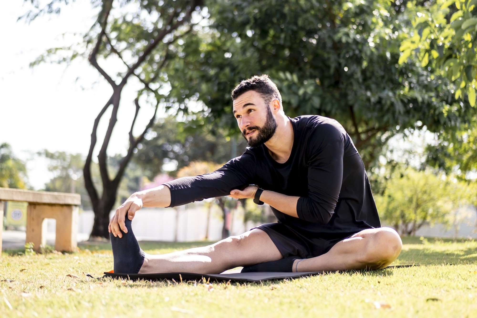 Young man smiling and sitting down in a park while stretching his right hamstring