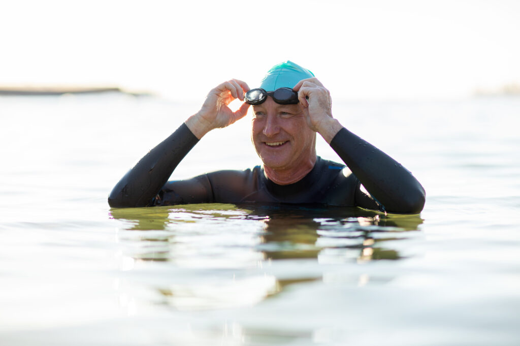 Man wearing goggles, a swim cap and a wetsuit smiling and relaxing in the water at the beach