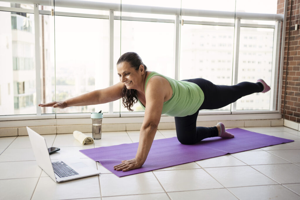 Woman smiling and watching her laptop while doing a pilates stretch in her apartment