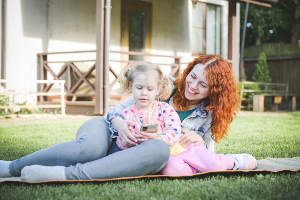 Woman smiling and lying down on the grass with her young daughter while looking at a phone
