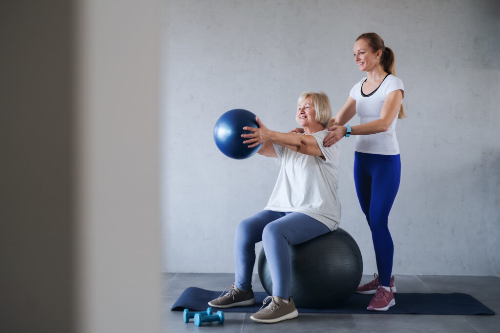 Woman sitting on fitball with another woman helping her with her workout