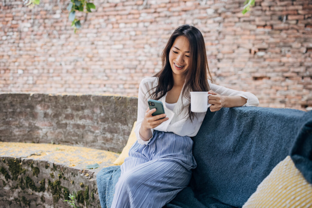 Woman sitting down outside on a brick seat, smiling and looking at phone with a cup in her left hand