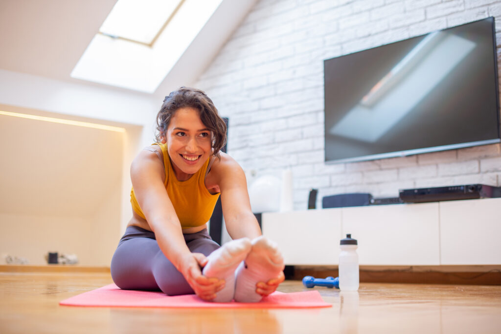 Woman in activewear sitting down inside and stretching with both hands touching her feet